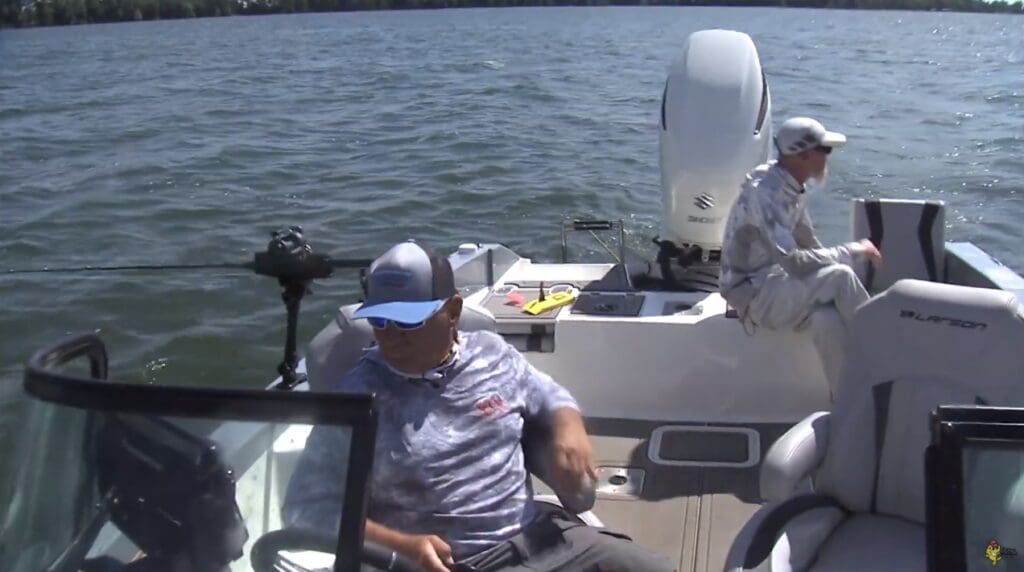Two people in fishing attire sit on a small motorboat on a lake. One, possibly following Mike Frisch’s tips, is at the back near fishing gear while the other sits near the front, both under a sunny sky with calm water.
