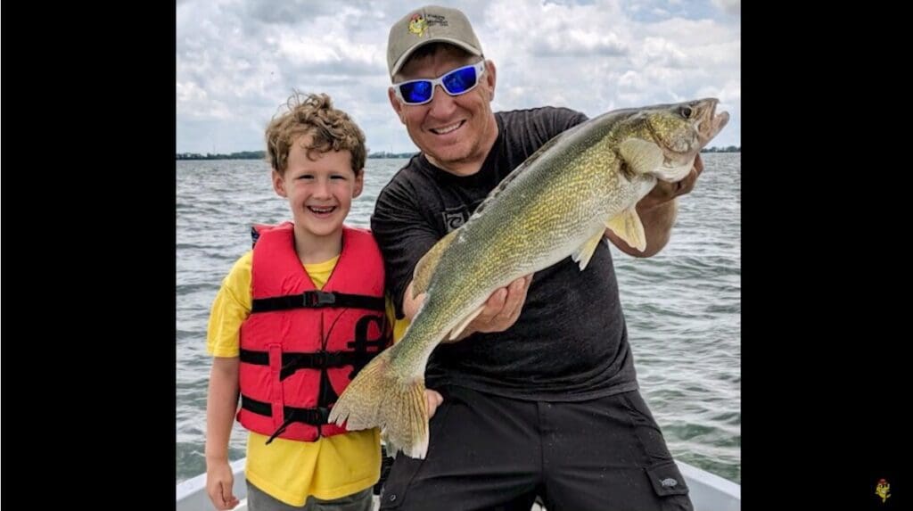A smiling adult wearing sunglasses and a cap holds a large fish after a successful fishing trip, standing next to a smiling child in a red life jacket on a boat, with water and a cloudy sky in the background.