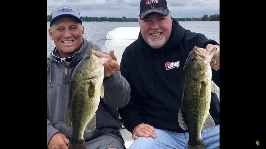 Two smiling men sit on a boat holding large bass fish, with a lake and cloudy sky in the background. Both wear caps and casual outdoor clothing, enjoying a successful fishing trip and appearing happy with their catch.
