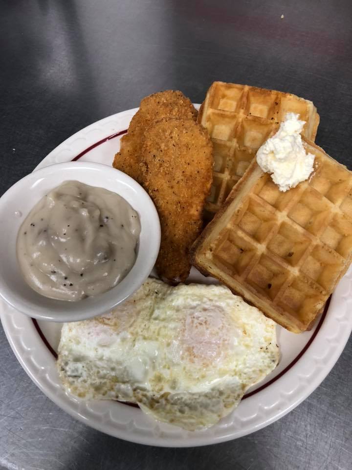 A plate at State Street Cafe featuring two waffles topped with a scoop of butter, two fried eggs, two breaded chicken tenders, and a small bowl of creamy sausage gravy.