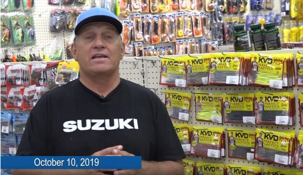 A man in a black SUZUKI shirt and blue cap stands in a fishing tackle store, surrounded by packs of fishing lures and gear. A blue bar on the bottom left displays the date October 10, 2019.