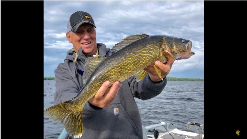 A person in a gray hoodie and cap, possibly Mike Frisch, smiles while holding a large yellow-green fish with sharp fins on a boat, with a lake and cloudy sky in the background.