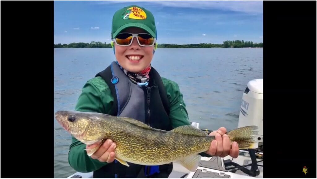 A smiling child wearing sunglasses and a green cap holds a large fish on a boat after a fun day of fishing, with a lake and trees in the background under a blue sky.