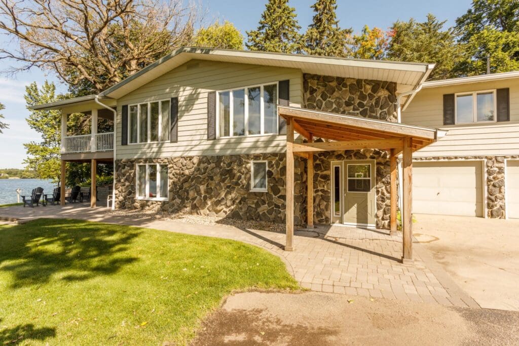 A two-story graystone house with wood siding, large windows, a covered entryway, attached garage, and a patio overlooking a lake, surrounded by trees and green grass under a clear sky.
