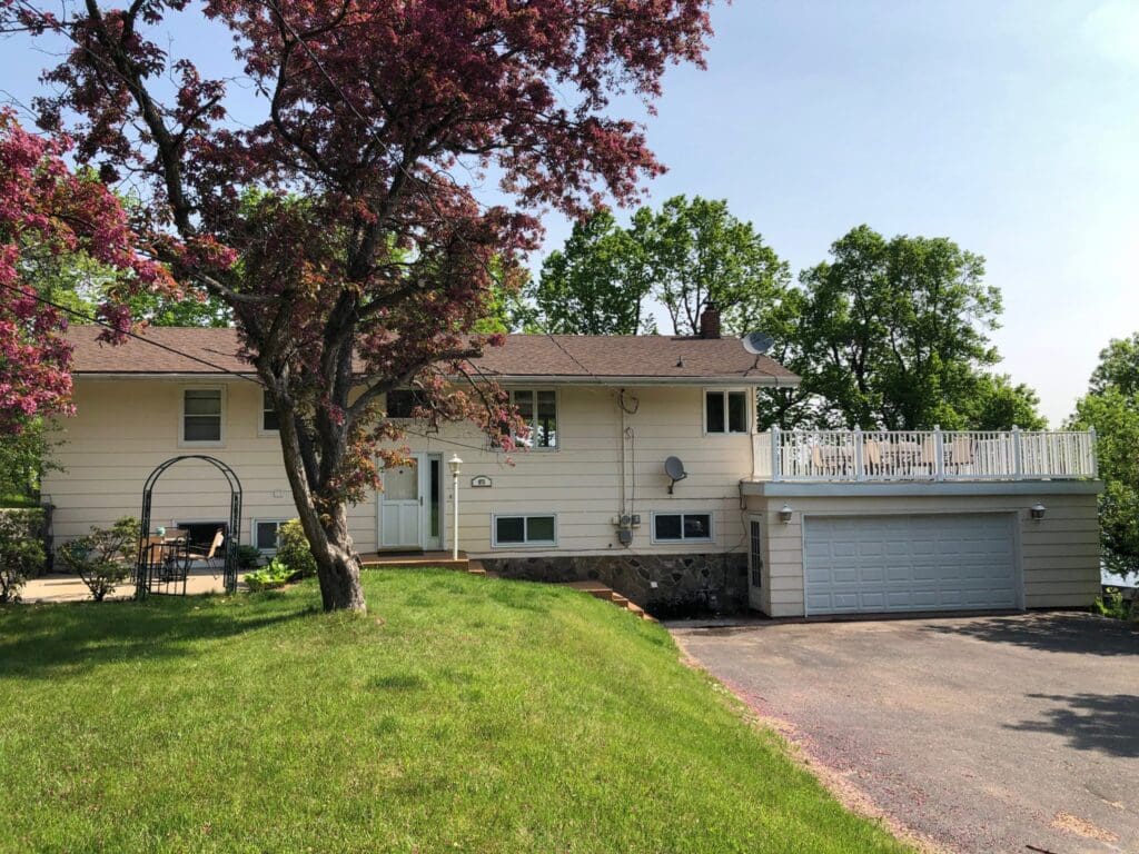 A beige split-level house with a two-car garage, white balcony railing, and a satellite dish. A large tree with red leaves stands out front—green grass surrounds the drive. Memories are Made on Brophy Shores in this inviting home.