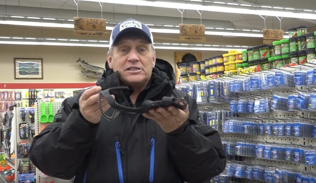 A man in a cap and jacket stands in a store aisle holding up a black fishing tool, with colorful fishing gear and supplies displayed on shelves behind him, reminiscent of expert angler Mike Frisch exploring top fishing picks.