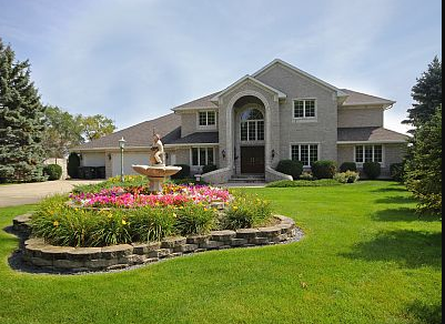 Lake Pointe Lodge is a large, two-story brick house with an arched entryway, surrounded by a well-maintained green lawn. A circular flower bed with a stone fountain sits in front, while trees gracefully frame the yard.