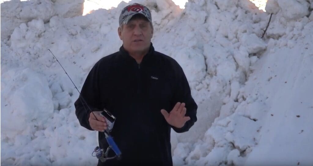 A man wearing a black jacket and a baseball cap stands in front of a large snowbank, holding a fishing rod in one hand and gesturing with the other, resembling angler Mike Frisch.