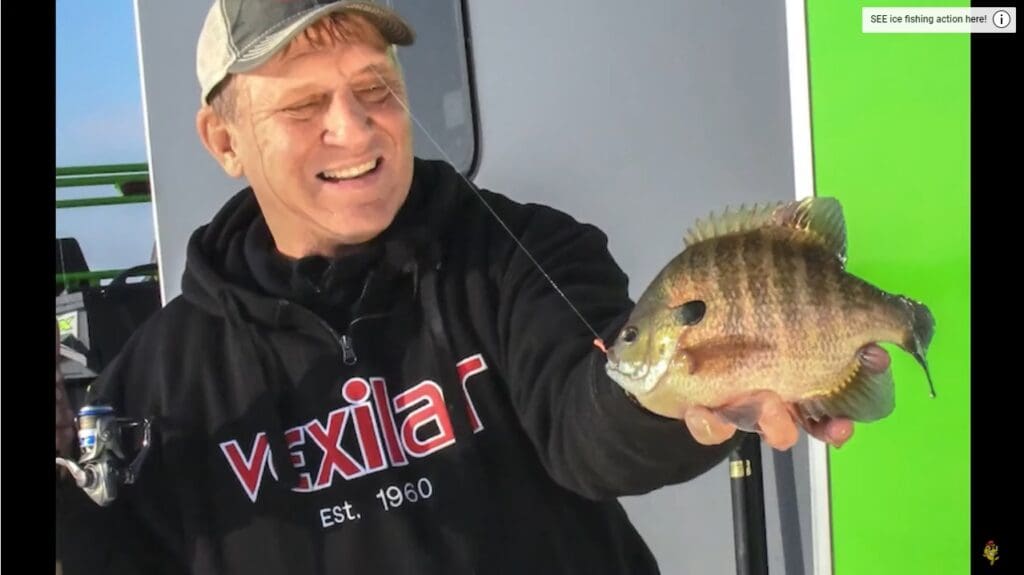 A smiling man, mike frisch, wearing a black "Vexilar" hoodie and cap, holds up a freshly caught fish on a fishing line, standing in front of a white and green background.