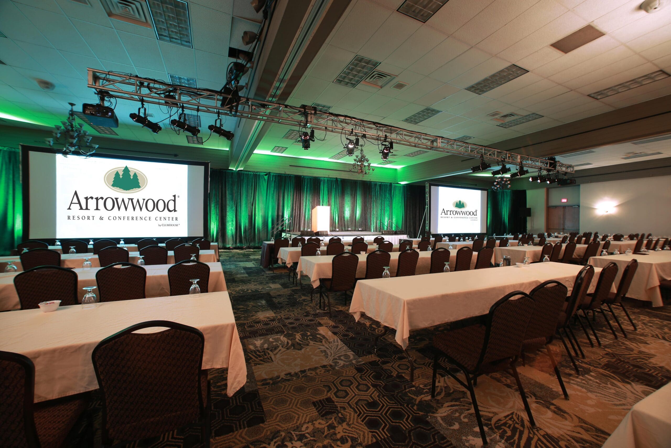 A large conference room at Arrowwood Resort features rows of tables and chairs facing two projection screens displaying the Arrowwood Resort & Conference Center logo. Green lighting highlights the stage and curtains at the front.