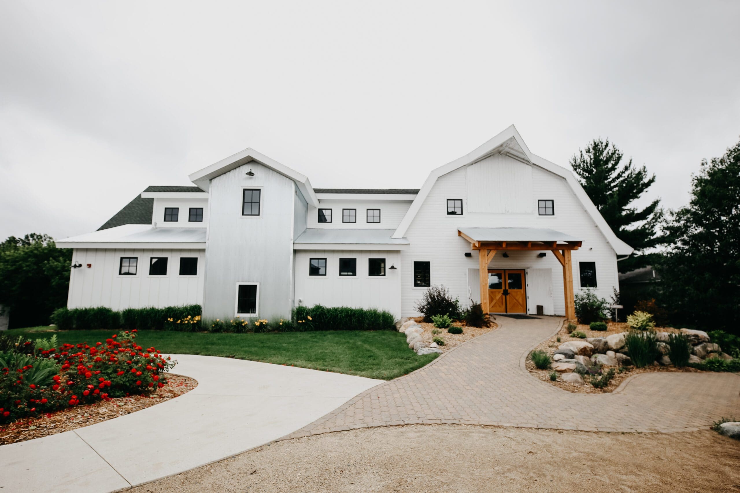 A large white modern barn-style building, Gathered Oaks features multiple gables and windows, surrounded by green grass, colorful flowerbeds, and a stone pathway leading to the entrance.