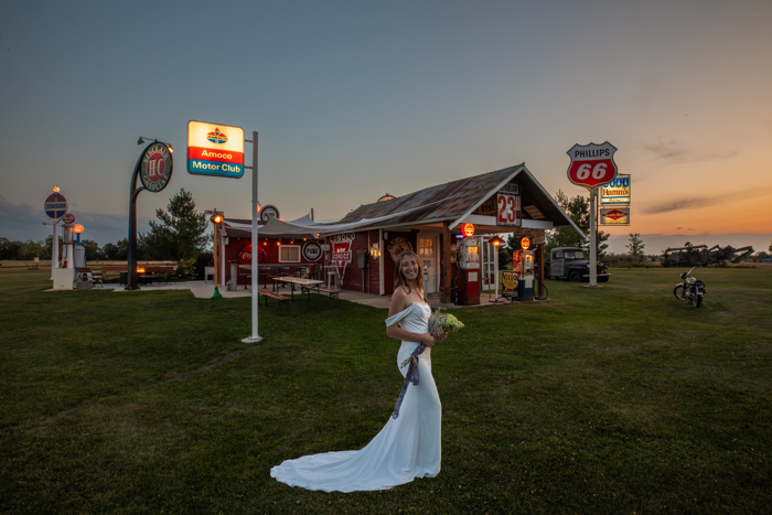 A bride in a white gown stands on grass at Adler Ranch in front of a vintage-style gas station at sunset. The red building, retro signs, and classic pumps set a nostalgic, Americana atmosphere.