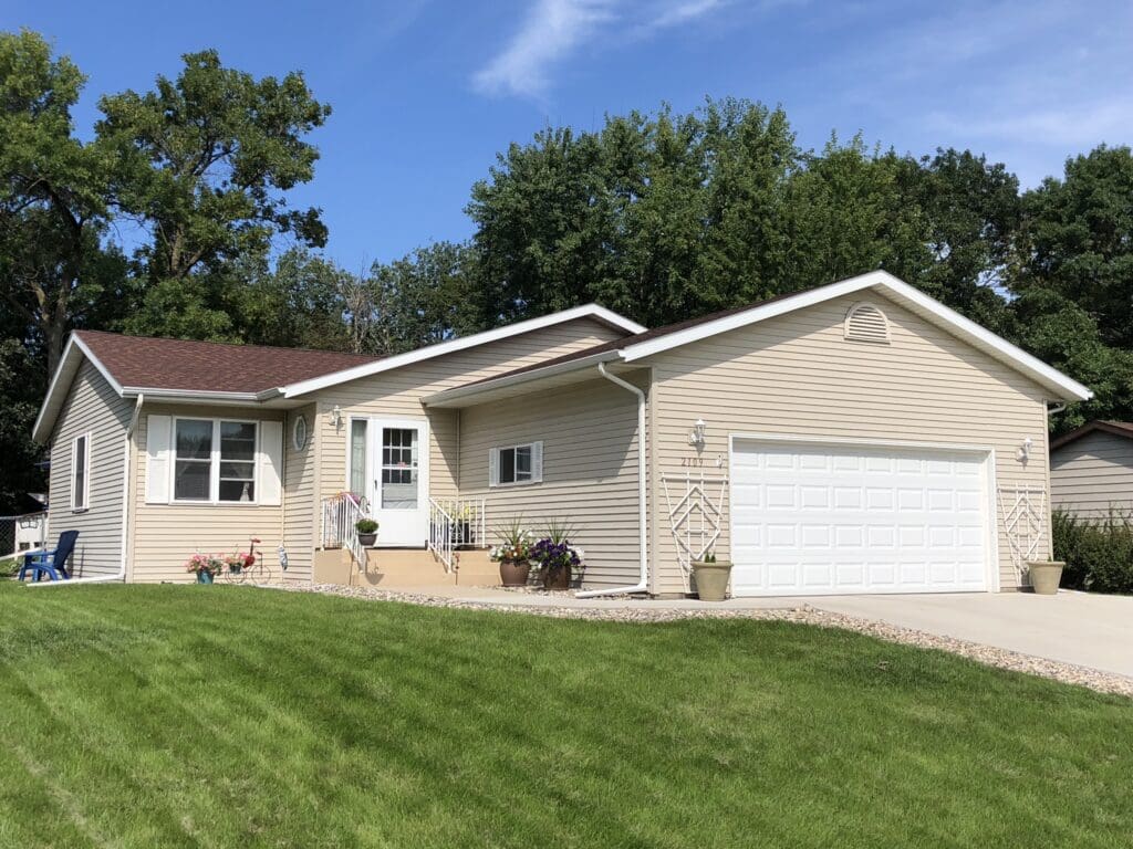Into the Blue Vacation Home is a single-story beige house with a red-brown roof, white garage door, front porch with railings, potted plants, and a well-kept green lawn, set against trees and a blue sky.