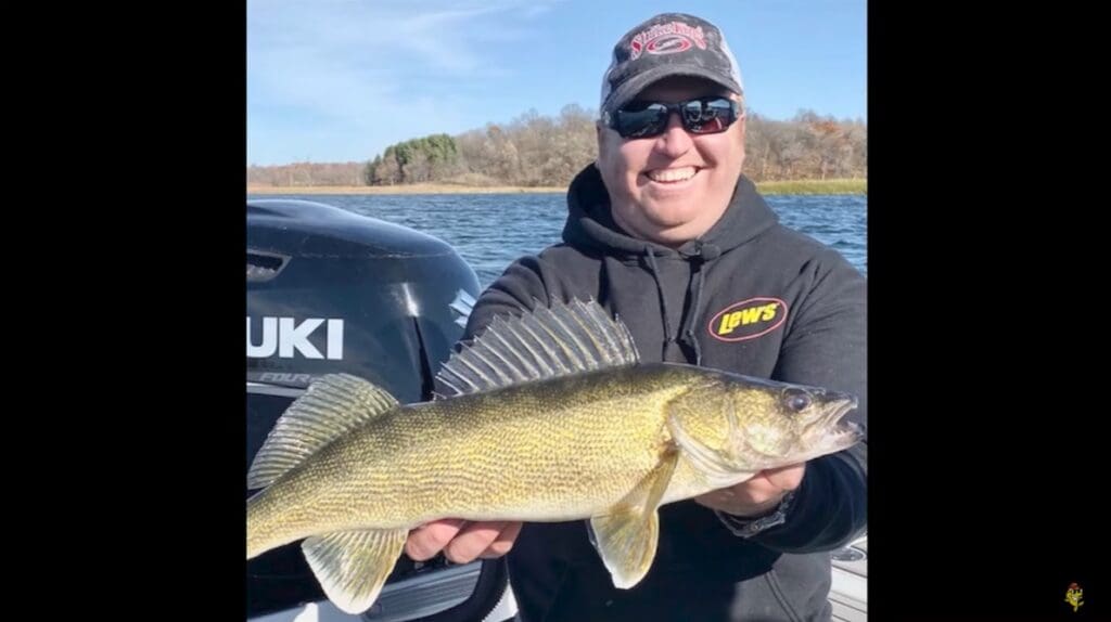 A smiling person wearing sunglasses and a black hoodie holds a large walleye fish on a boat, with a Suzuki motor and a lake in the background—just another great day of fishing with Mike Frisch.