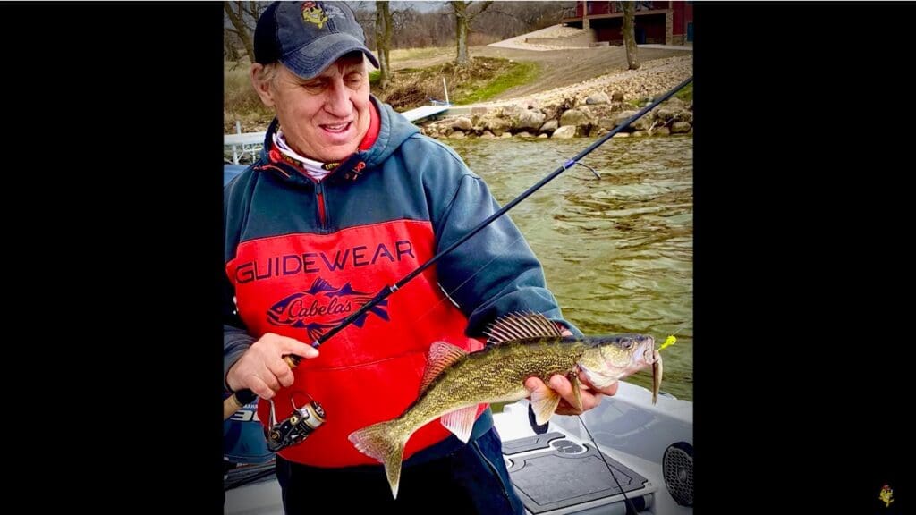 A man in a red and navy jacket, possibly Mike Frisch, holds a fishing rod and proudly displays a freshly caught fish while standing on a boat in a lake near a rocky shoreline.