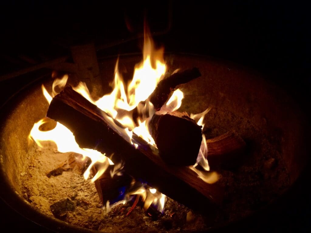 A close-up of a campfire at night in the Lakes Area, with glowing orange flames rising from burning logs arranged in a fire pit, surrounded by ash and embers—perfect for socially distant summer activities.
