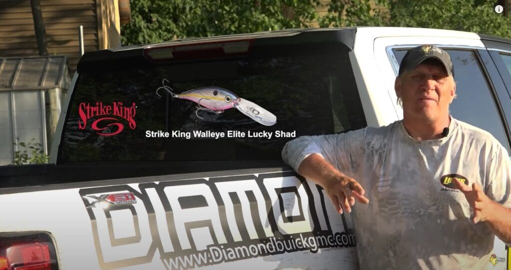 A man in a cap and camo shirt stands by a white truck with "DIAMOND" and a web address on the tailgate. The rear window displays a Strike King fishing lure ad, a large fishing lure image, and the name Mike Frisch.