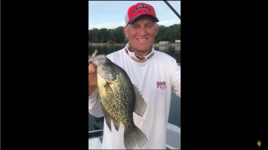 A man in a white long-sleeve shirt and red cap smiles while holding up a large speckled fish on a boat, with water and trees in the background—capturing a classic Mike Frisch fishing moment.