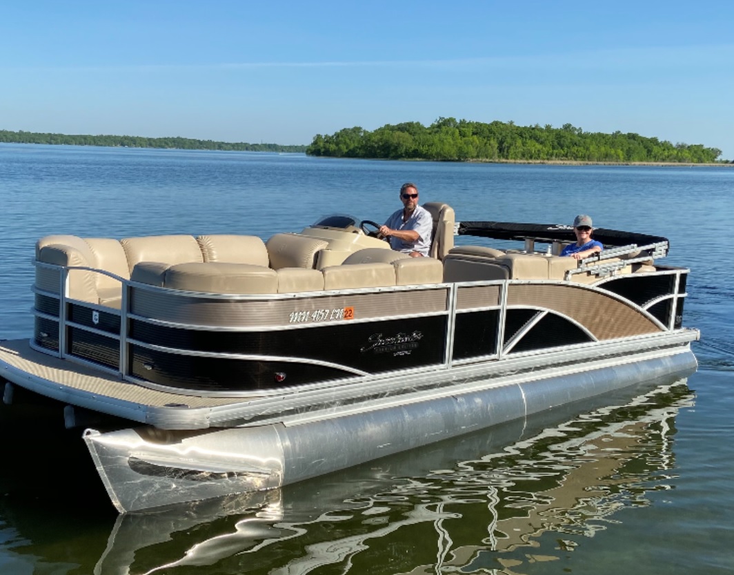 A man and a child ride a beige and black pontoon boat on calm water, with a small green island and clear blue sky in the background—another perfect outing from Toon Tastic Pontoon Rentals.