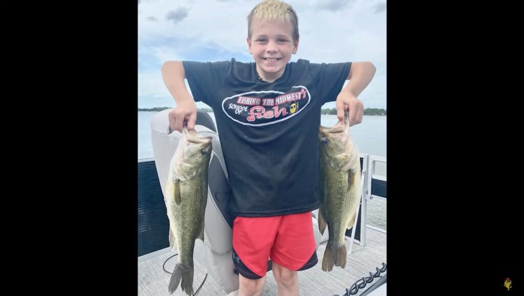 A smiling boy stands on a boat, proudly holding up two large fish—one in each hand—after a successful day of fishing. He wears a black T-shirt and red shorts, with water and sky stretching out behind him.