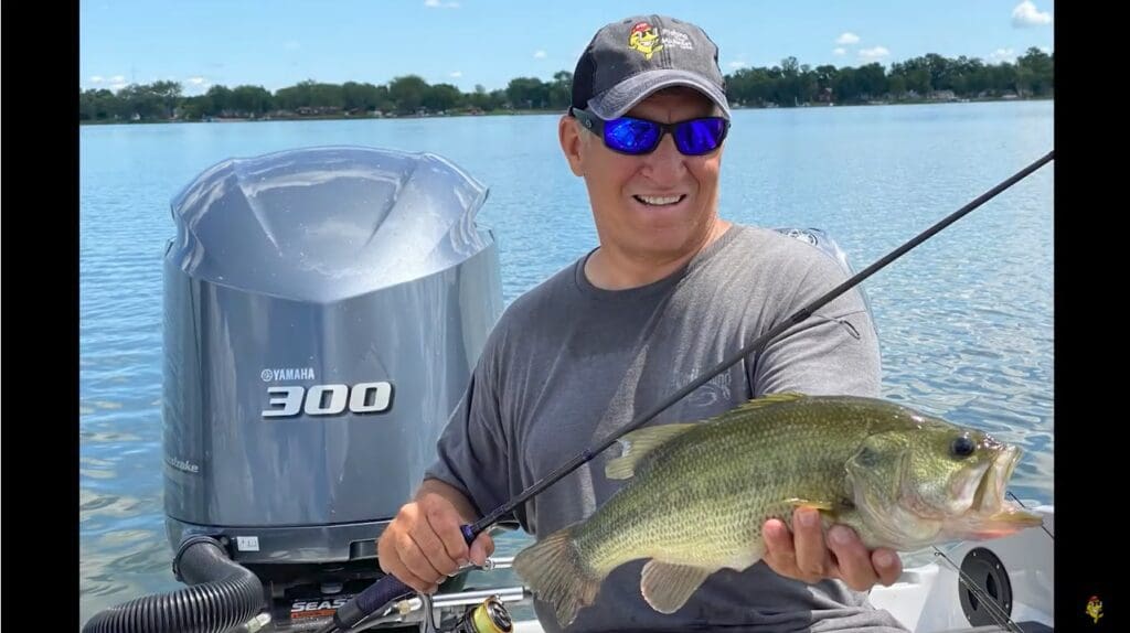 A man wearing sunglasses and a cap smiles while fishing, holding a large fish on a boat in a lake, with a Yamaha 300 outboard motor behind him and trees in the background.