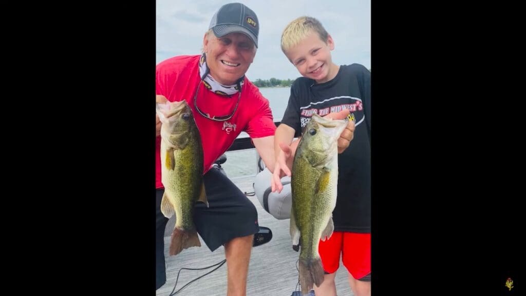 An adult and a smiling boy stand on a dock holding large fish they caught while fishing, with a lake and cloudy sky in the background. Both appear happy and proud of their catches.