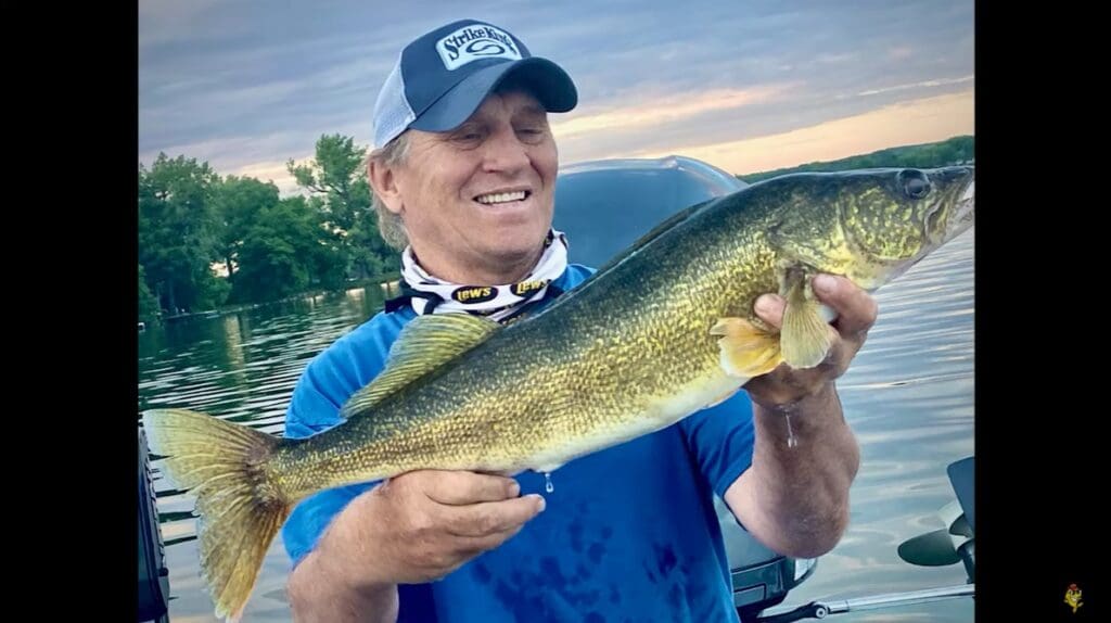A smiling person in a blue shirt and cap shows off their fishing skills, holding a large fish with greenish-yellow scales on a boat, with water and trees in the background during sunset.
