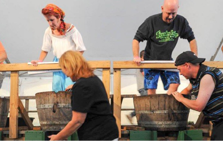 Two men and a woman grapple grape stomping in wooden barrels on a raised platform at an Alexandria event. Another woman stands in front, slightly blurred, as they enjoy one of the area’s favorite autumn activities.