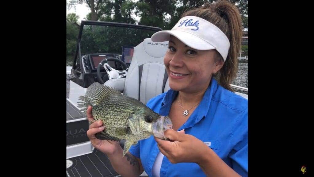 A smiling woman in a blue shirt and white visor holds a large fish beside a boat on the water, with trees in the background—just like an angler from Mike Frisch’s fishing adventures.