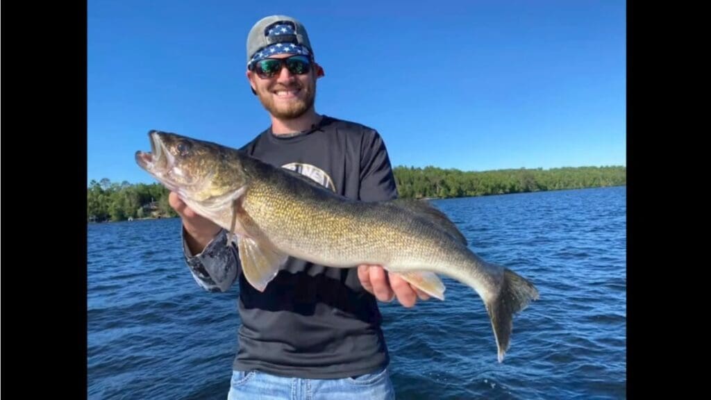 A smiling man, Mike Frisch, wearing sunglasses and a cap, holds a large fish on a boat, with blue water and a forested shoreline in the background under a clear blue sky.