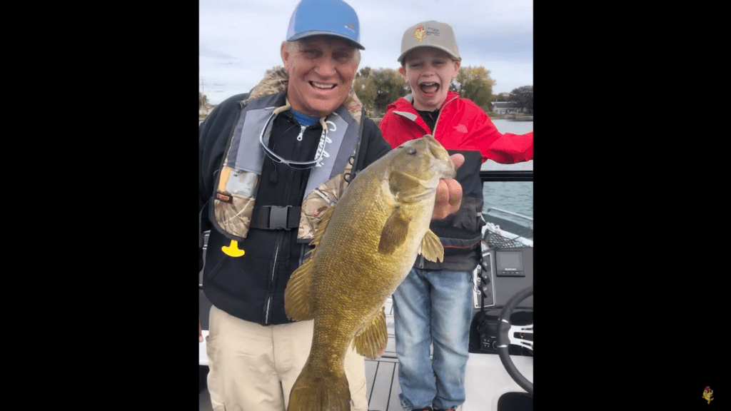 An older man and a young boy smile excitedly on a boat, proudly holding up a large fish they caught while fishing. The boy wears a red jacket and cap, the man sports a blue cap and life vest, with water and trees in the background.