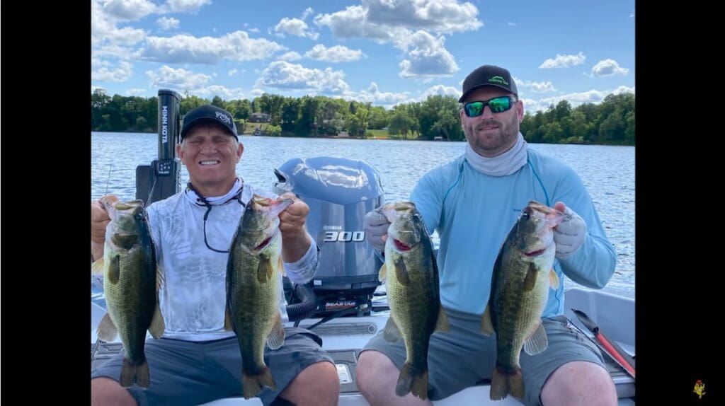 Two men, including Mike Frisch, sit on a boat holding up large bass they caught, smiling at the camera. They are on a lake surrounded by trees and set against a blue sky with scattered clouds.