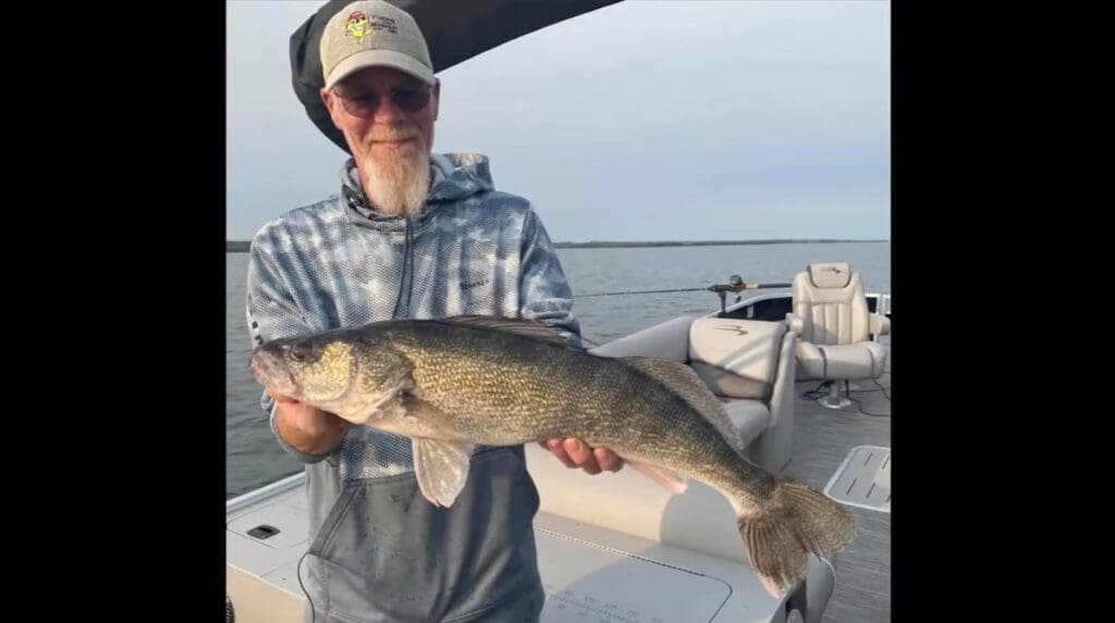 A man wearing glasses, a cap, and a blue hoodie stands on a boat holding a large fish with both hands. The water and sky are visible in the background, capturing the excitement of Mike Frisch's fishing adventure.