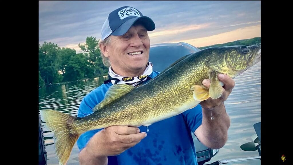 A smiling man in a blue shirt and cap holds a large, golden-brown fish, proudly showing off his fishing catch by the lake, with trees and a cloudy sky in the background.