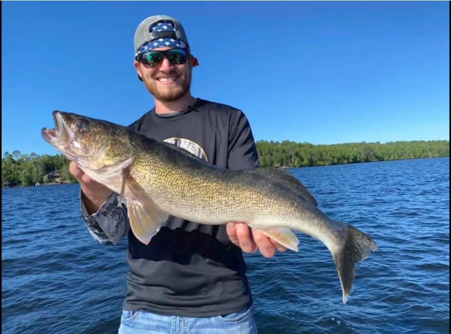 A smiling man wearing sunglasses and a cap shows off his catch while fishing on a boat in a lake, with trees and a clear blue sky in the background.
