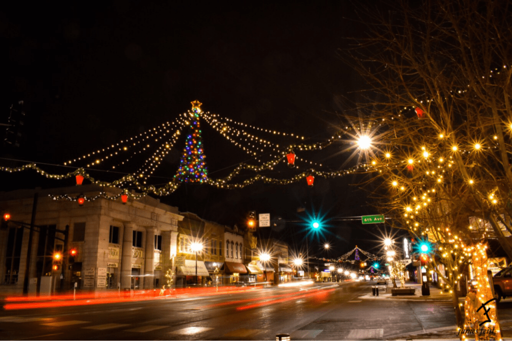 A downtown street at night is decorated with Christmas lights, including a lit tree and string lights above, as storefronts glow for holidays shopping and car light trails line the road—reminding everyone to shop safe this season.