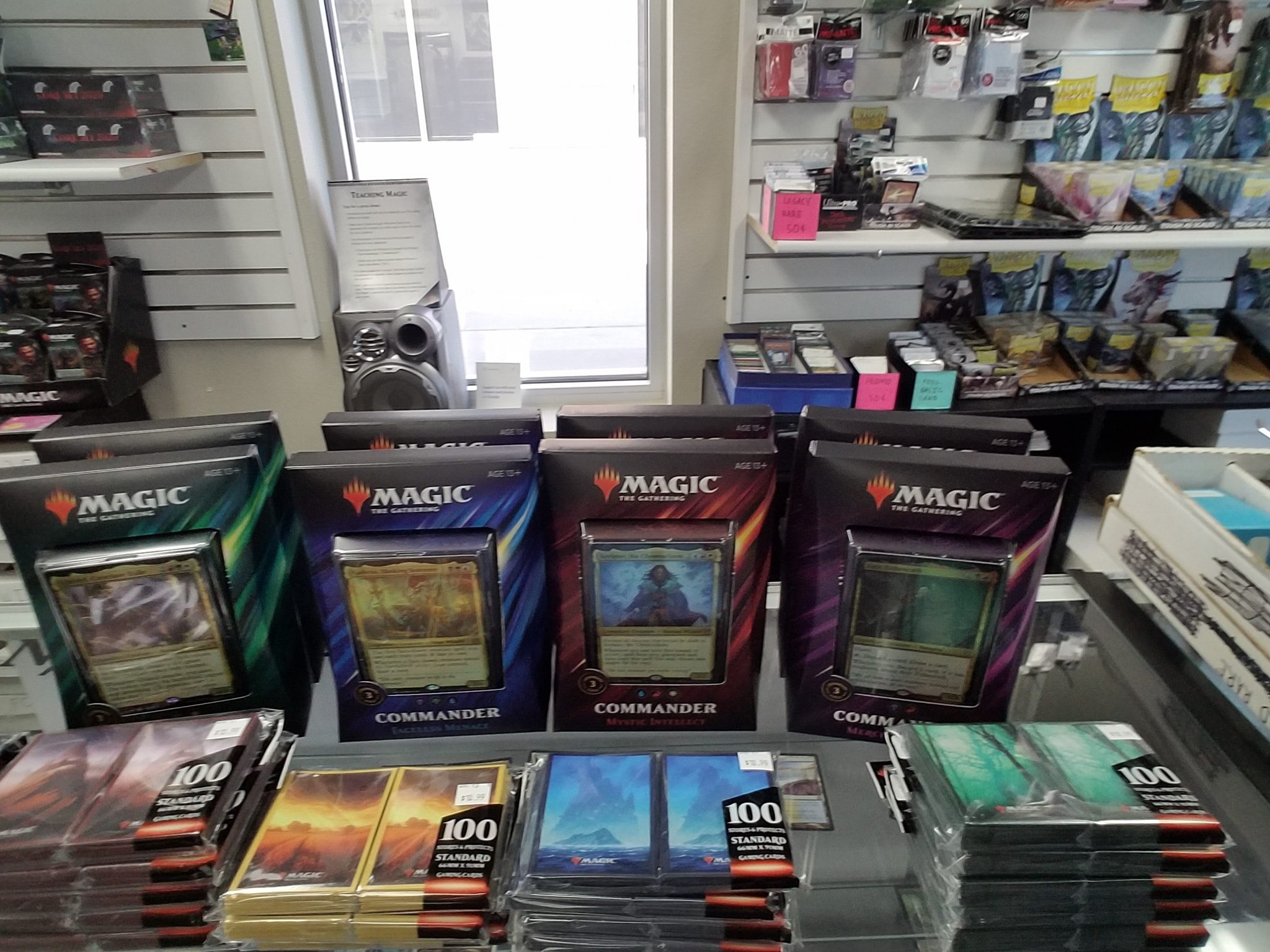 Four Magic: The Gathering Commander decks are displayed on a glass counter at Elemental Games, surrounded by card sleeves and packs, with shelves of gaming products visible in the background.