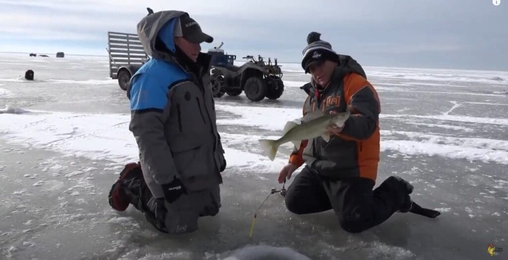 Two people in winter gear kneel on a frozen lake; one, possibly Mike Frisch, holds a fish and a fishing rod. An ATV and sled are in the background, with ice fishing holes visible on the icy surface.
