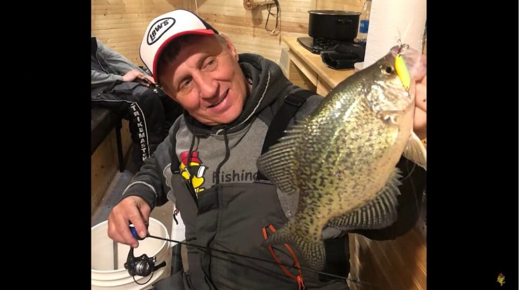 A smiling man in fishing gear, possibly Mike Frisch, holds up a large crappie fish indoors, sitting on a white bucket with a fishing rod in his other hand.