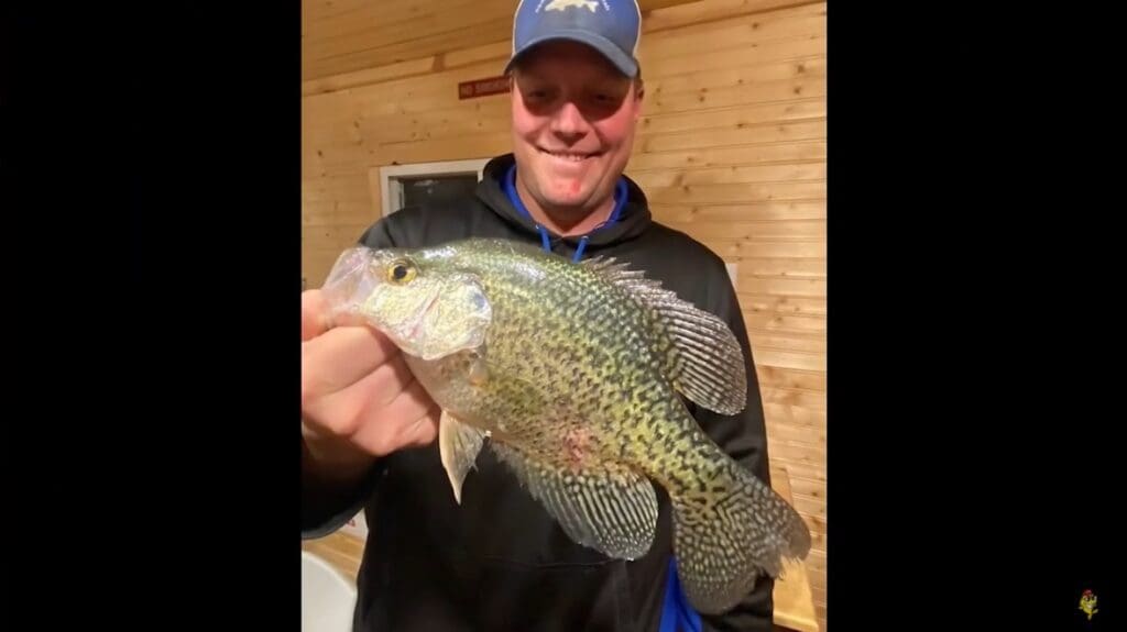 A smiling man in a blue cap and black hoodie, identified as Mike Frisch, holds a large, spotted crappie fish indoors against a wooden wall background.