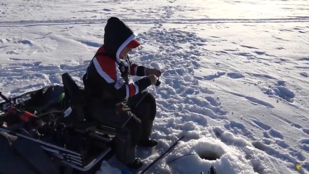 A person sits on a folding chair ice fishing, dressed in winter clothing with a hood, much like mike frisch. They hold a fishing rod over a hole in the snow-covered ice, surrounded by equipment and footprints.