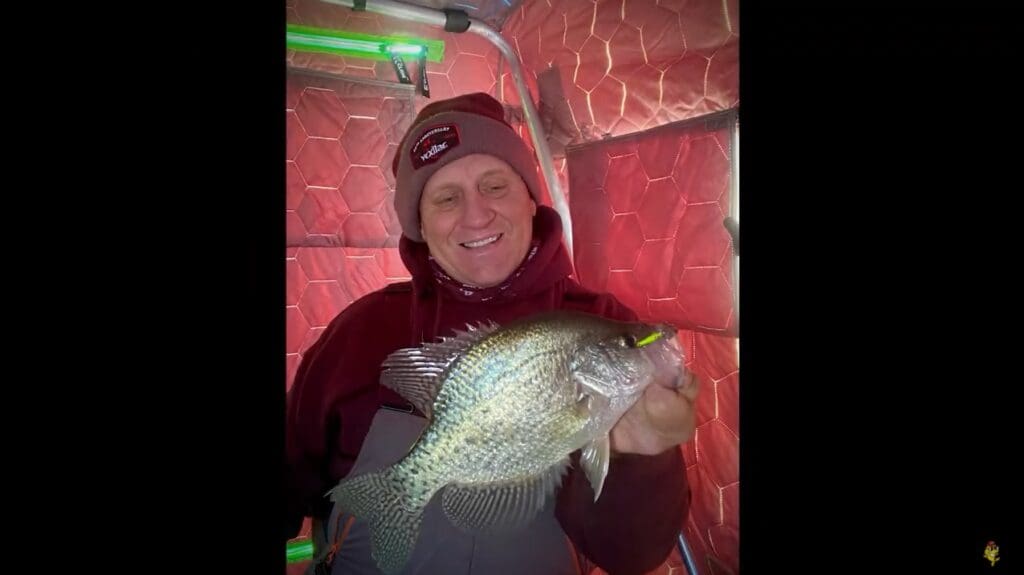 Mike Frisch, in a maroon hoodie and beanie, smiles while holding a large fish inside an ice fishing shelter with red, hexagonal-patterned walls.