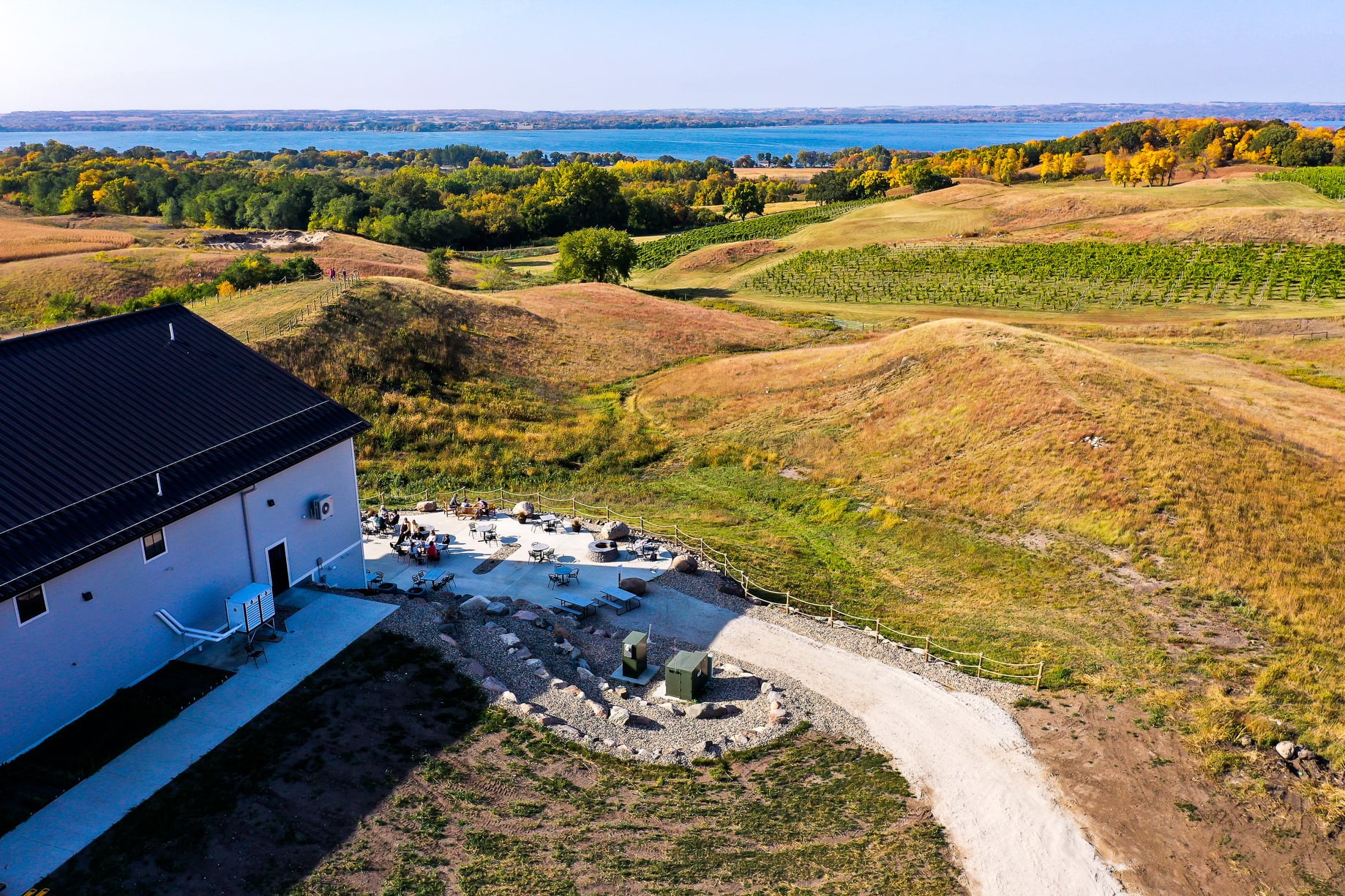 A white building sits beside a patio with tables and people at Rolling Forks Vineyards, surrounded by grassy hills and a gravel path, overlooking a river and tree-lined landscape under a clear blue sky.