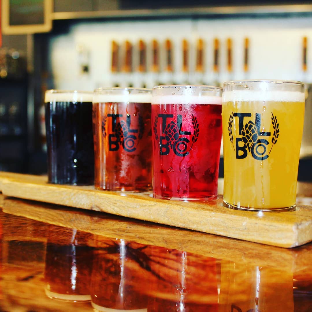 Four glasses of beer in varying hues, from dark brown to light yellow, are arranged on a wooden paddle atop a polished bar counter at Thousand Lakes Brewing, with beer taps softly blurred in the background.