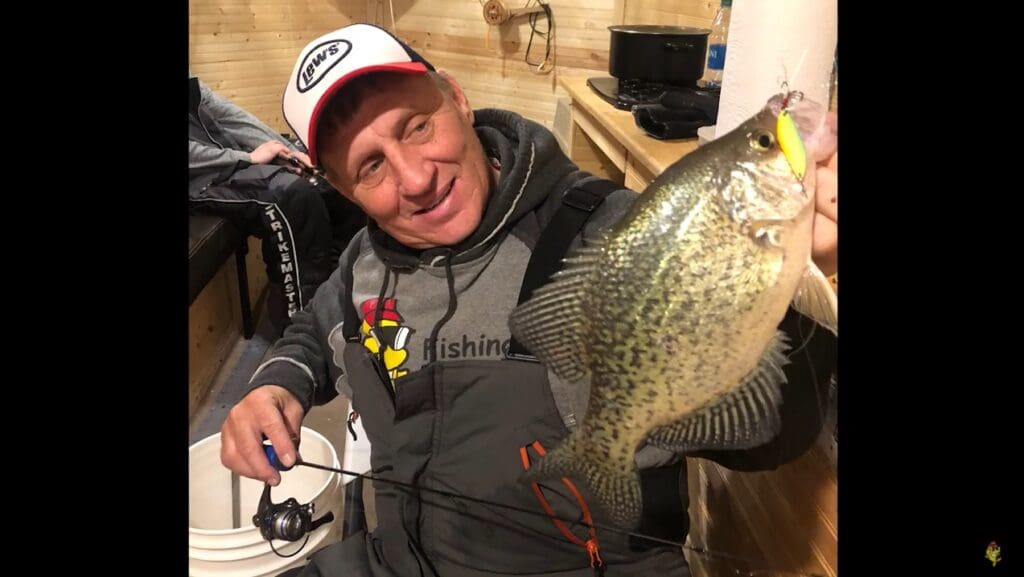 A smiling man, mike frisch, in fishing gear holds up a large speckled fish inside a wooden cabin, showing it to the camera. He has a fishing rod in one hand and wears a white cap with logos.