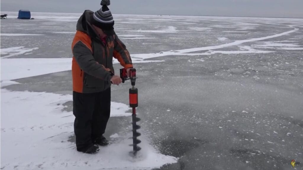 A person dressed in winter clothing uses an auger to drill a fishing hole in the ice on a frozen lake, with snow and ice covering the surface and a blue tent visible in the distance.