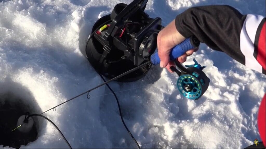 A person, possibly named Mike, ice fishing holds a blue and black fishing rod over a hole in the ice, with a black fishing reel device and equipment on the snowy surface nearby.