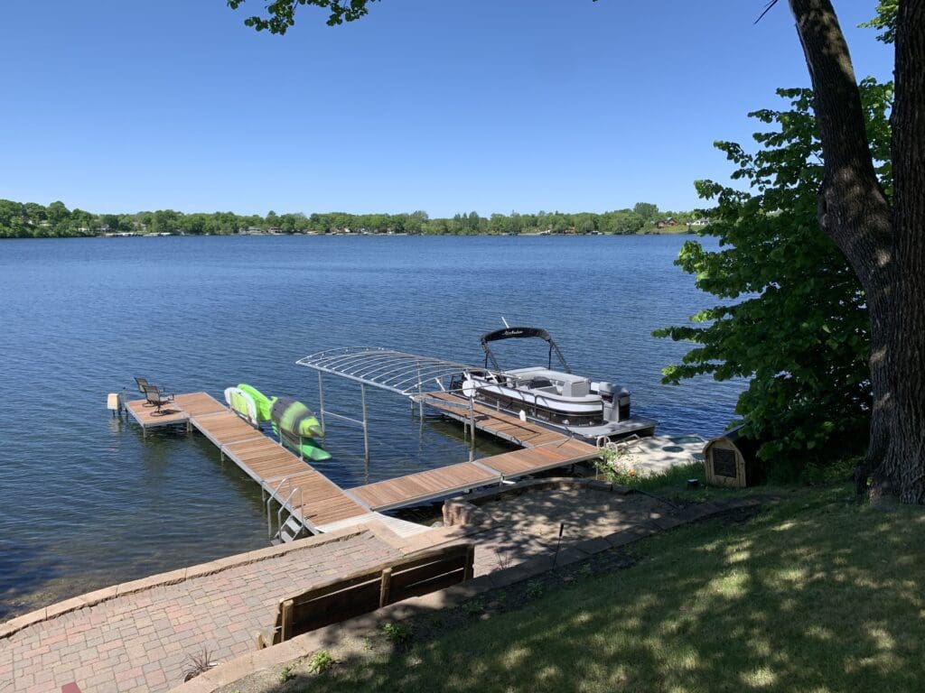 A peaceful lakeside scene at Lake Cowdry Retreat, with a wooden dock, pontoon boat, two green kayaks, and a bench on a grassy hill under a tree, all overlooking calm blue water and distant green trees beneath a clear sky.