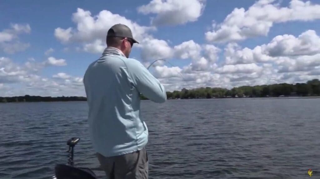 A person in a light blue shirt and cap stands on a boat, fishing on a lake under a partly cloudy sky, with calm water and a treeline visible in the distance—enjoying open water instead of ice fishing conditions.