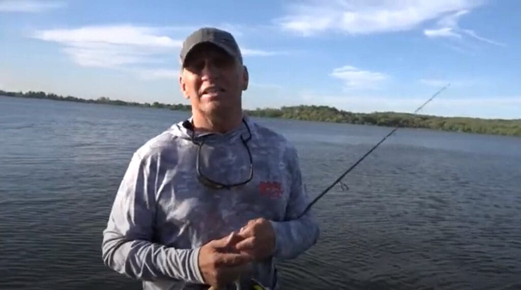 A man in a cap and sunglasses rests a fishing rod while standing by a large lake under a blue sky with scattered clouds, hoping to catch crappies. Trees and shoreline are visible in the distance.